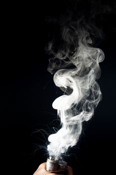 Male hand holds vape without atomiser. Column of vape clouds splash from coil on black background. Fog is white. Stock isolated white smoke with spray boiling glycerine.