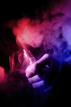 Male hand holds vape with atomiser in thick vape fog or smoke. Vape clouds around hand with vape on black background. Fog is dual color — blue and red. Stock isolated colorful smoke with spray glycerine.