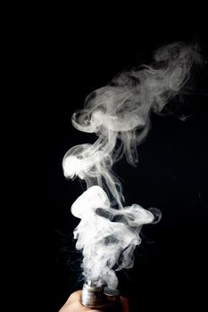 Male hand holds vape without atomiser. Column of vape clouds splash from coil on black background. Fog is white. Stock isolated white smoke with spray boiling glycerine.