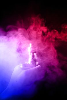 Male hand holds vape with atomiser in thick vape fog or smoke. Vape clouds around hand with vape on black background. Fog is dual color — blue and red. Stock isolated colorful smoke with spray glycerine.