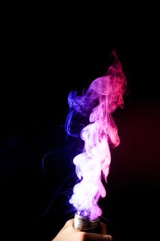 Male hand holds vape. Vape clouds splash from vape coil on black background. Fog is dual color — blue and red. Stock isolated dual colour smoke with spray boiling glycerine.