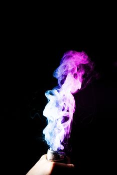 Young man holds vape in his hand. Vape clouds splash from vape coil on black background. Fog is dual color — blue and purple. Stock isolated colorful smoke with spray boiling glycerine.