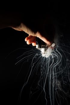 Male hand holds vape upside down. Vape clouds splash from coil on black background. Fog is white. Stock isolated white smoke with spray boiling glycerine.