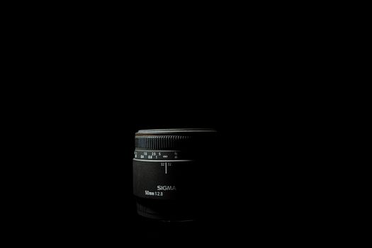 Ukraine; Kyiv; 20.02.2019 Sigma 50mm f1:2,8 DG EX Macro lens. Underexposed photo of DSLR camera lens lying on black background. Stock photo isolated. Low light photo and large depth of field.