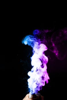 Young man holds vape in his hand. Vape clouds splash from vape coil on black background. Fog is dual color — blue and purple. Stock isolated colorful smoke with spray boiling glycerine.