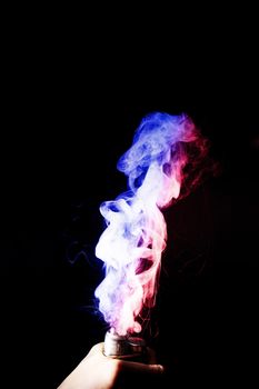 Male hand holds vape. Vape clouds splash from vape coil on black background. Fog is dual color — blue and red. Stock isolated dual colour smoke with spray boiling glycerine.