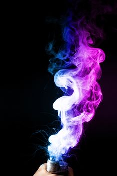 Male hand holds vape. Vape clouds splash from vape coil on black background. Fog is dual color — blue and purple. Stock isolated colorful smoke with spray boiling glycerine.