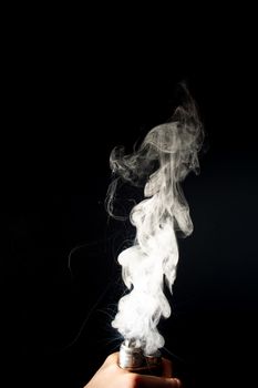 Man hand holds vape. Vape clouds splash from coil on black background. Fog is white. Stock isolated white smoke with spray boiling glycerine around.