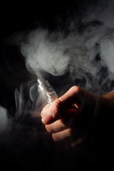 Male hand holds vape in thick vape fog or smoke. Vape clouds around hand with vape on black background. Fog is white. Stock isolated white smoke.