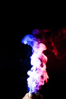 Male hand holds vape. Vape clouds splash from vape coil on black background. Fog is dual color — blue and red. Stock isolated dual colour smoke with spray boiling glycerine.