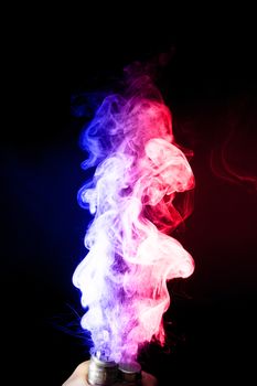 Male hand holds vape. Vape clouds splash from vape coil on black background. Fog is dual color — blue and red. Stock isolated dual colour smoke with spray boiling glycerine.