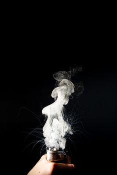 Man hand holds vape. Vape clouds splash from coil on black background. Fog is white. Stock isolated white smoke with spray boiling glycerine around.