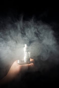 Male hand holds vape in thick vape fog or smoke. Vape clouds around hand with vape on black background. Fog is white. Stock isolated white smoke.