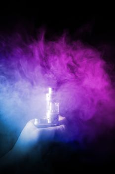 Male hand holds vape in thick vape fog or smoke. Vape clouds around hand with vape on black background. Fog is dual color — blue and purple. Stock isolated colorful smoke with spray glycerine.