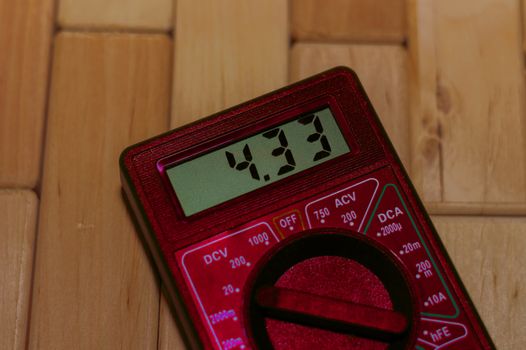 Red digital measuring multimeter on wooden floor. It shows 4.33V or fully charged battery. Includes voltmeter, ampermeter, ohmmeter.