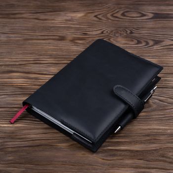 Black handmade leather notebook cover with notebook and pen inside on wooden background. Stock photo of luxury business accessories.
