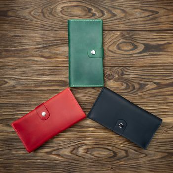 Three handmade leather wallets lies on textured wooden backgroud. Green, red and black wallets up to down view.