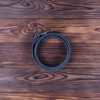 Black color handmade belt lies on textured wooden background. The belt is twisted into a ring. Up to down view. Stock photo of businessman accessories.