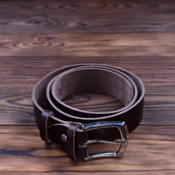 Brown color handmade belt lies on textured wooden background. The belt is twisted into a ring. Closeup side view. Stock photo of businessman accessories.