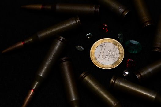Rifle ammo with a lot of gemstones around one euro coin on black background. Symbolizes the war for money and one of the world's problems.