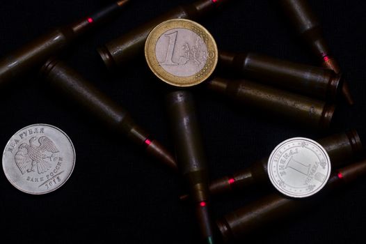 One euro, one Russian ruble and Ukrainian hryvna coins with rifle military ammo on black background. Symbolizes war for money- biggest problem in world.