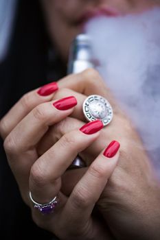 Caucasian woman with red nails manicure and antique ring on finger holds small vape. Smoking alternative vay. Woman exhales thick smoke. Life without cigarettes. Woman-vaper. Small e-cigarette.