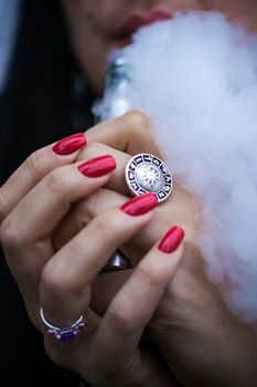 Caucasian woman with red nails manicure and antique ring on finger holds small vape. Smoking alternative vay. Woman exhales thick smoke. Life without cigarettes. Woman-vaper. Small e-cigarette.