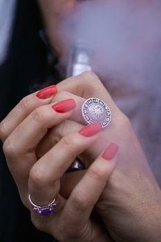 Caucasian woman with red nails manicure and antique ring on finger holds small vape. Smoking alternative vay. Woman exhales thick smoke. Life without cigarettes. Woman-vaper. Small e-cigarette.