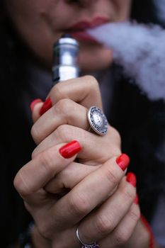 Caucasian woman with red nails manicure and antique ring on finger holds small vape. Smoking alternative vay. Woman exhales thick smoke. Life without cigarettes. Woman-vaper. Small e-cigarette.