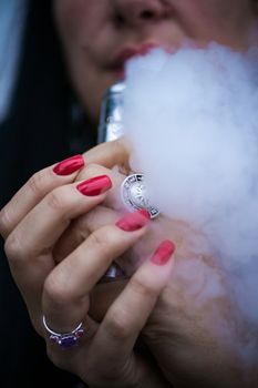 Caucasian woman with red nails manicure and antique ring on finger holds small vape. Smoking alternative vay. Woman exhales thick smoke. Life without cigarettes. Woman-vaper. Small e-cigarette.