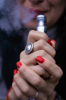 Caucasian woman with red nails manicure and antique ring on finger holds small vape. Smoking alternative vay. Woman exhales thick smoke. Life without cigarettes. Woman-vaper. Small e-cigarette.