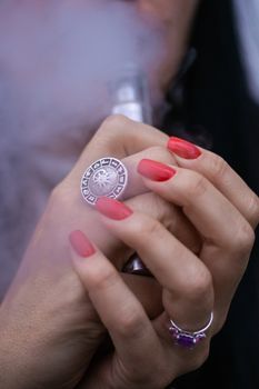 Caucasian woman with red nails manicure and antique ring on finger holds small vape. Smoking alternative vay. Woman exhales thick smoke. Life without cigarettes. Woman-vaper. Small e-cigarette.