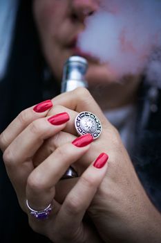 Caucasian woman with red nails manicure and antique ring on finger holds small vape. Smoking alternative vay. Woman exhales thick smoke. Life without cigarettes. Woman-vaper. Small e-cigarette.