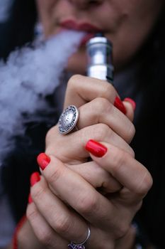 Caucasian woman with red nails manicure and antique ring on finger holds small vape. Smoking alternative vay. Woman exhales thick smoke. Life without cigarettes. Woman-vaper. Small e-cigarette.