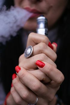 Caucasian woman with red nails manicure and antique ring on finger holds small vape. Smoking alternative vay. Woman exhales thick smoke. Life without cigarettes. Woman-vaper. Small e-cigarette.