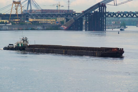 A large ship sails along a wide river in an industrial zone. The industrial zone and bridge are located in the background.