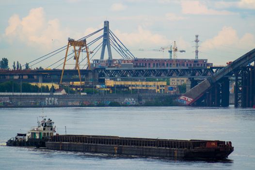 A large ship sails along a wide river in an industrial zone. The industrial zone and bridge are located in the background.