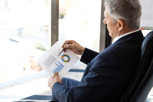 Mature businessman sitting alone in office with large panoramic windows with view at city and looking at diagrams and financial reports