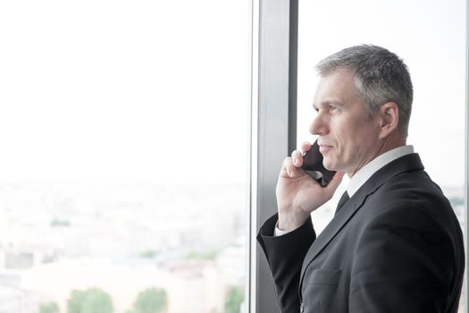 Mature businessman talking on a mobile phone standing by the window with view on city
