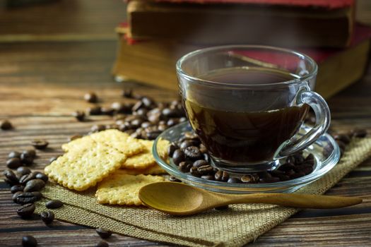 Black coffee in clear glass cup and coffee beans with crackers and old book on wooden table.