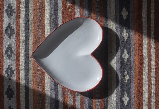 Heart shaped white plate with red edge on rustic rug with shadows decorative background.