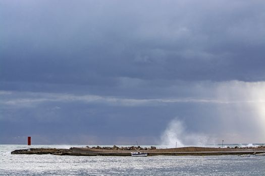 Mediterranean sea view with pier against dark skies with dramatic light on splashing wave on a winter day in Mallorca, Spain.