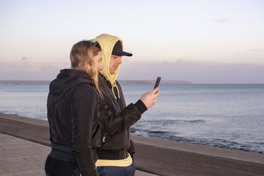 Handsome young natural and casual looking couple with hood jackets black and yellow watch sunset and photographing with smartphone on a beach.