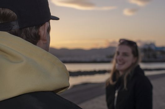 Over the shoulder shot of guy in yellow hood jacket to smiling woman blurry, casual looking couple with hood jackets watch sunset in Majorca, Spain.
