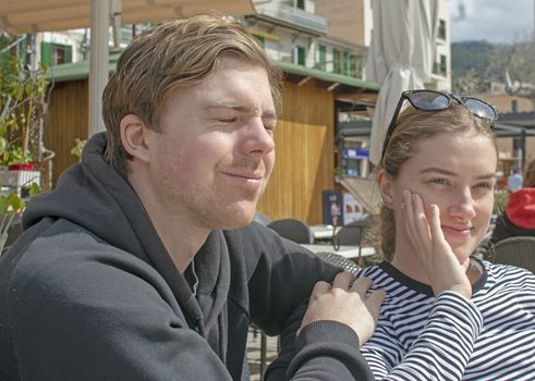 Young handsome happy couple sit close in cafe and squint in stark sunshine on a sunny spring day in Mallorca, Spain.