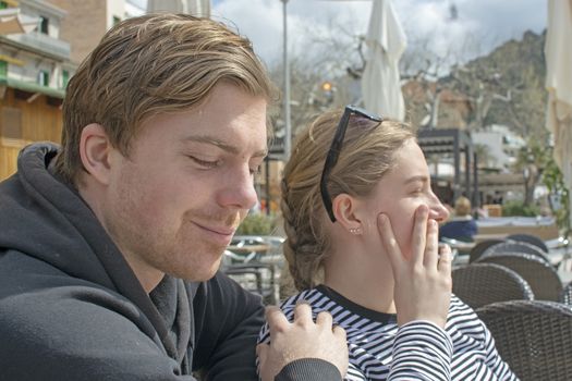 Young handsome happy couple sit close in cafe and squint in stark sunshine on a sunny spring day in Mallorca, Spain.