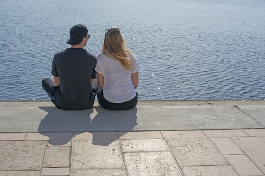 Young couple sit in sunshine watch water glitter on stone pier on a spring day in Mallorca, Spain.