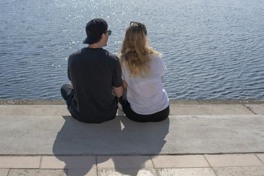 Young couple sit in sunshine watch water glitter on stone pier on a spring day in Mallorca, Spain.