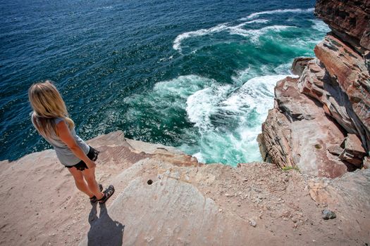 A female takes in the ocean views from a cliff top ledge that is unfenced. Beautiful colours in teh shallower water