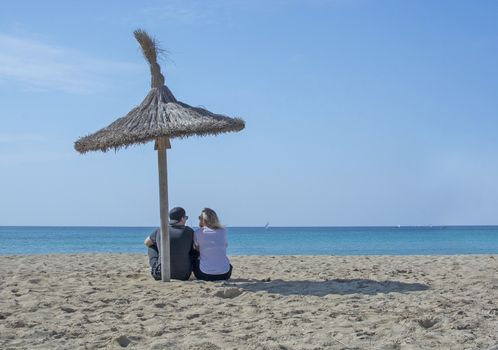 Young natural and casual sporty couple sit on beach under parasol and watch the ocean horizon in Mallorca, Spain.